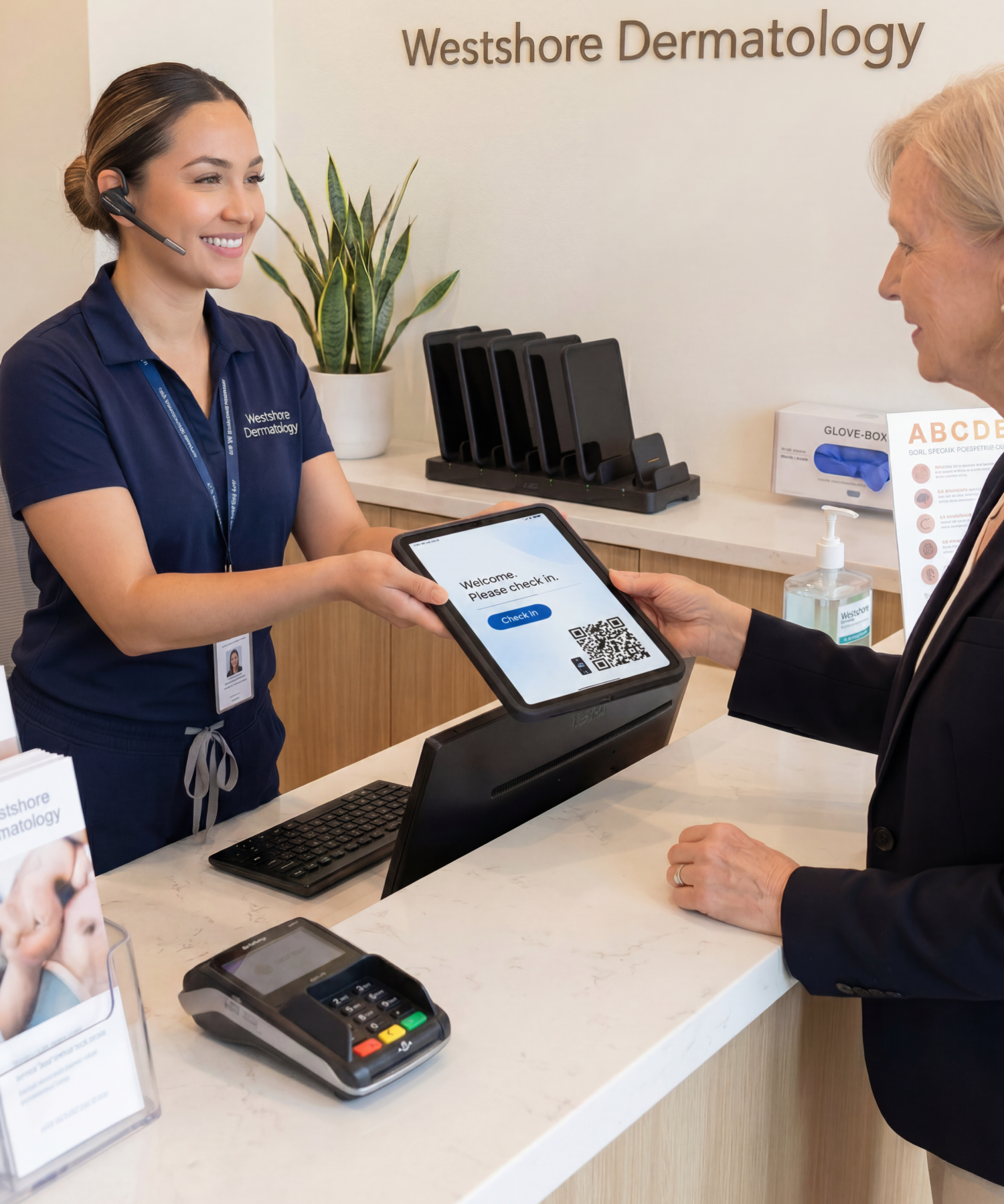 Dermatology receptionist handing a patient a CONNECT Case iPad for check-in, with a MultiDock 6 charging station and ABCDE skin-check poster in frame
